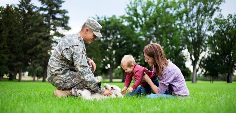 A man is dressed in a military outfit, petting his dog. His wife and small child are close by